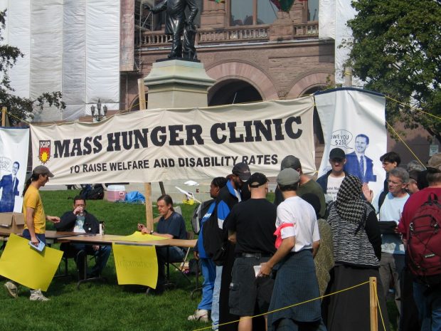 Ein grosses Banner mit den Wörtern „Mass Hunger Clinic to Raise Welfare and Disability Rates“ (Massen-Hungerklinik, um die Sozialhilfe- und Behindertenraten zu erhöhen) vor dem Parlament von Ontario. Zwei Menschen sitzen an einem Informationstisch. Andere Menschen stehen in einer Reihe oder sind in ein Gespräch verwickelt.