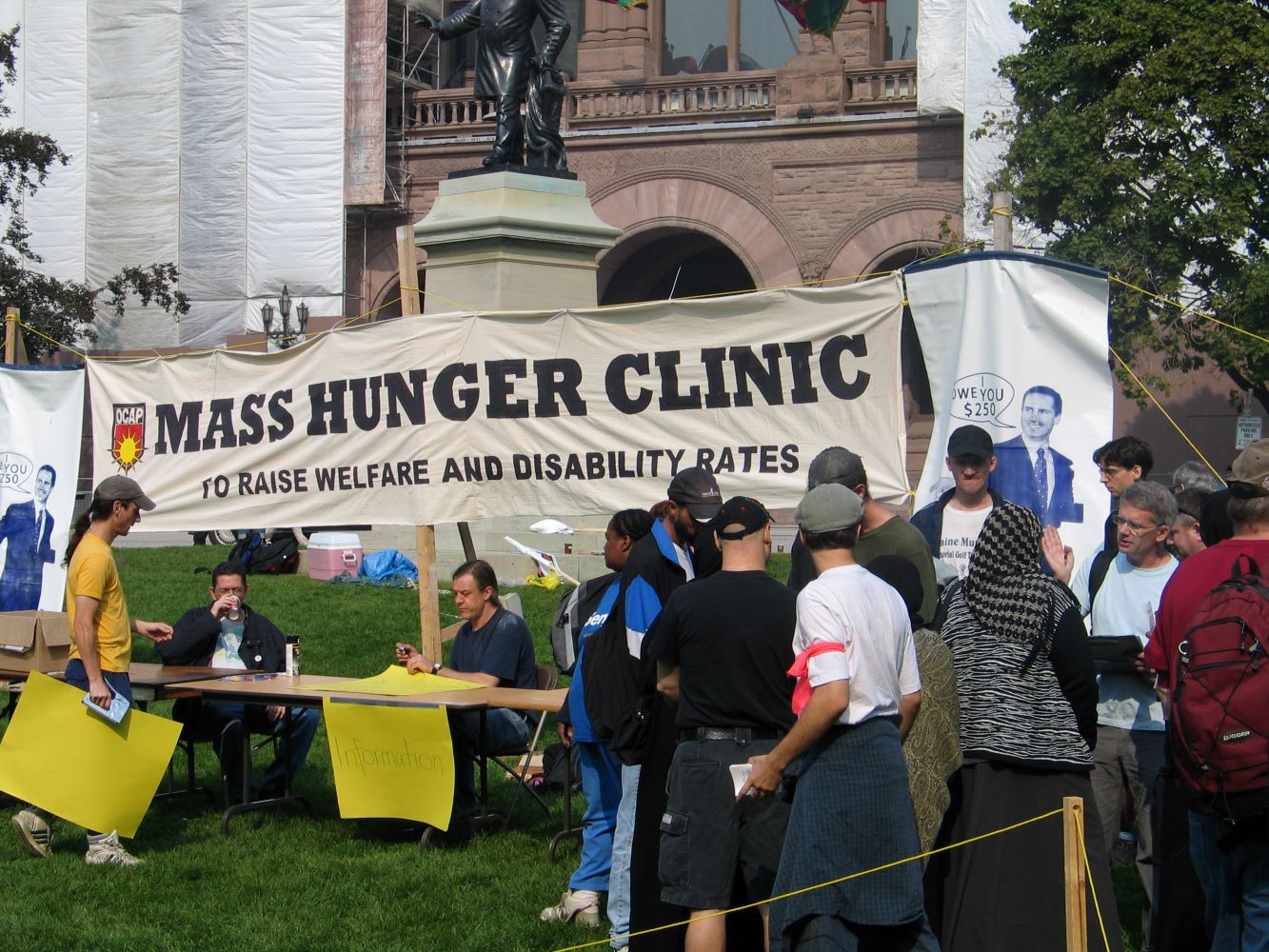 Ein grosses Banner mit den Wörtern „Mass Hunger Clinic to Raise Welfare and Disability Rates“ (Massen-Hungerklinik, um die Sozialhilfe- und Behindertenraten zu erhöhen) vor dem Parlament von Ontario. Zwei Menschen sitzen an einem Informationstisch. Andere Menschen stehen in einer Reihe oder sind in ein Gespräch verwickelt.