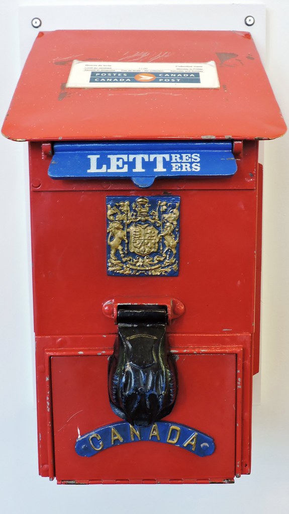 Ein rot-metallischer Briefkasten im alten Stil mit einem moderneren Logo der Canada Post obendrauf. Auf der Vorderseite befindet sich eine blaue Klappe mit der Aufschrift „Letters“ auf Englisch und „Lettres“ auf Französisch. Unter der Klappe ist eine kunstvolle Gravur eines Wappens, eines schwarzen Schlossdeckels und der Schrift: Kanada.