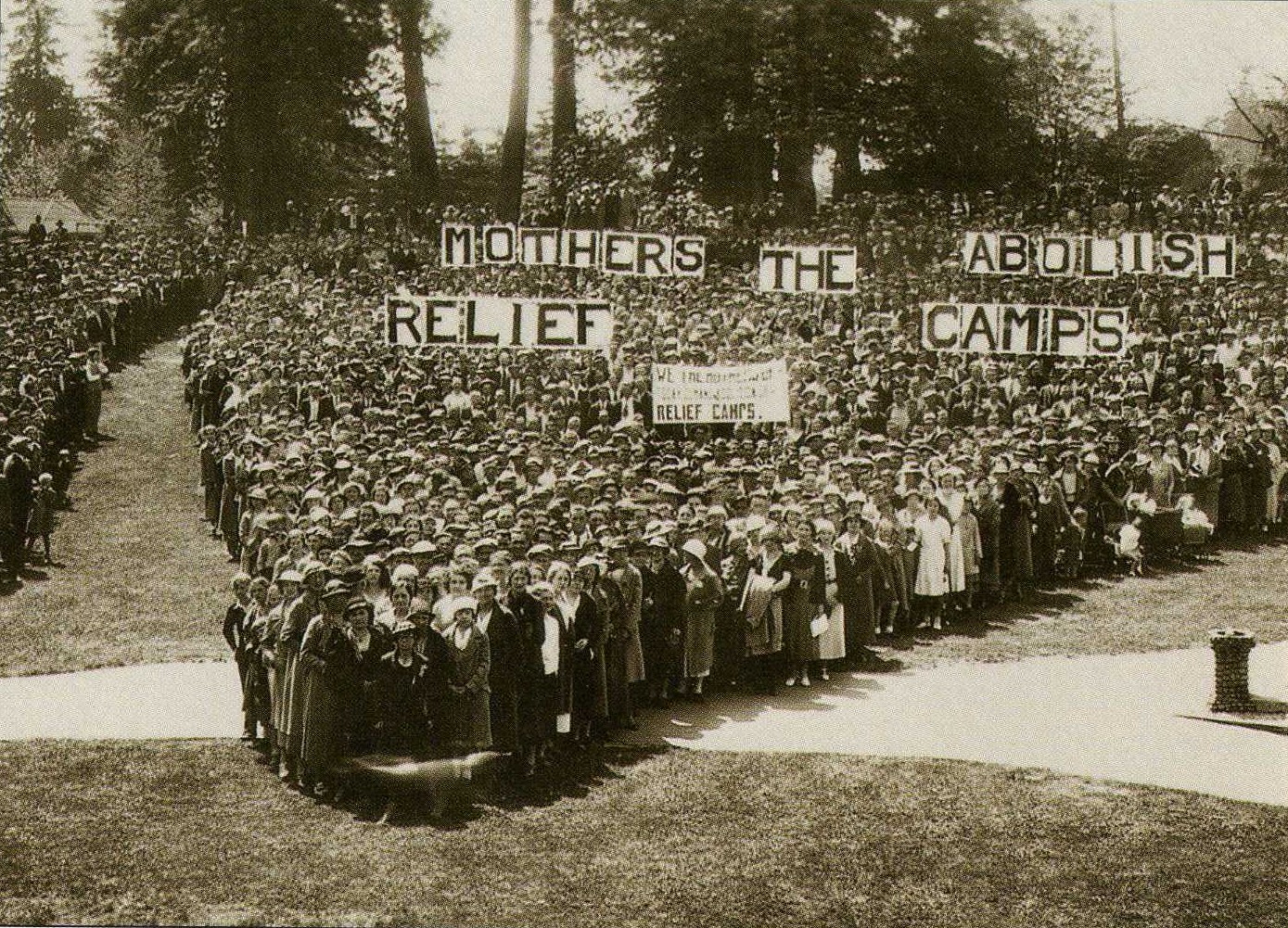 Une photographie en noir et blanc, prise depuis les airs, montre un groupe important de manifestantes, réunies, portant des banderoles sur lesquelles on peut lire : « Mothers Abolish The Relief Camps » (Mères, abolissons les camps de réfugiés)