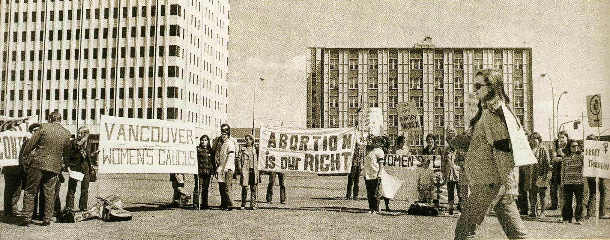 Eine Schwarz-Weiß-Fotografie, die eine Gruppe protestierender Frauen zeigt, die Beschriftungen und Banner tragen und vor zwei Gebäuden stehen. Eines der Banner besagt: „Abortion is Our Right“ (Abtreibung ist unser Recht) und das andere besagt: „Vancouver Women's Caucus“ (Vancouver Frauenfraktion). Eine Frau geht über den Platz vor der Gruppe.