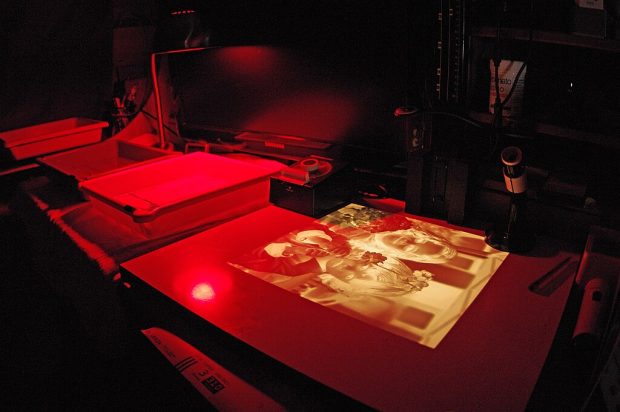 Eine fotografische Dunkelkammer, die von blassrotem Licht beleuchtet wird. Im Bild sind verschiedene Tabletts mit Flüssigkeiten sichtbar. Auf dem Tisch daneben liegen belichtete und trockene Fotos.