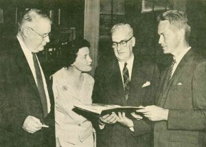 Photo en noir et blanc montrant trois hommes et une femme posant avec un livre.
