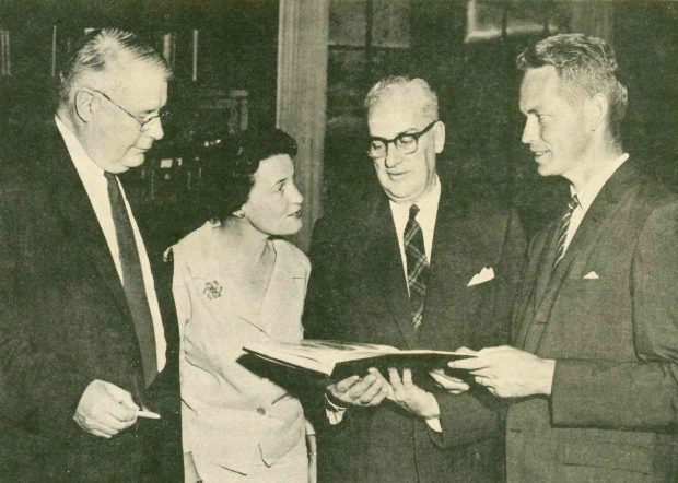 Black and white photo of three men and a woman posing with a book.