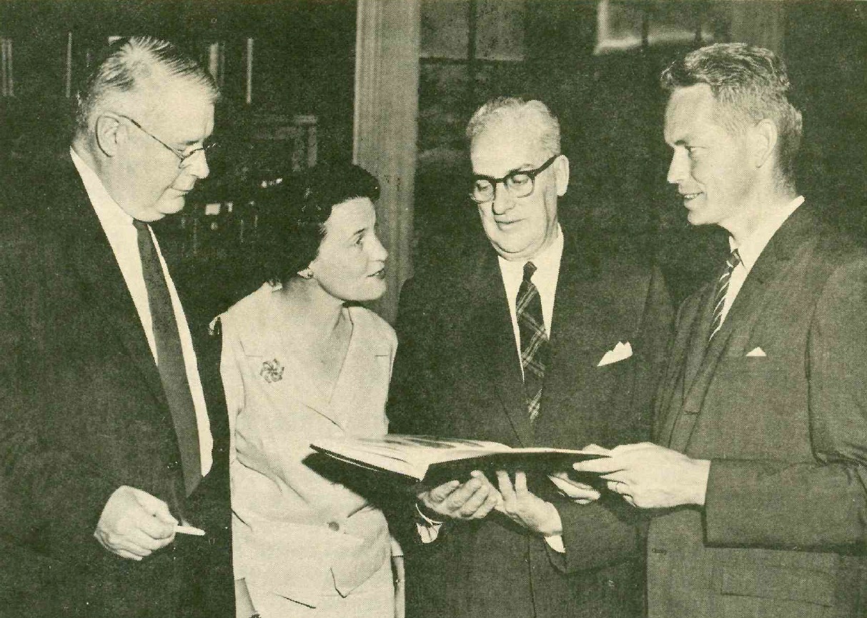 Black and white photo of three men and a woman posing with a book.