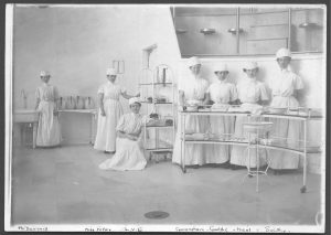 Black and white photo of seven nurses posing in an operating room wearing early 19th C white nursing uniforms.