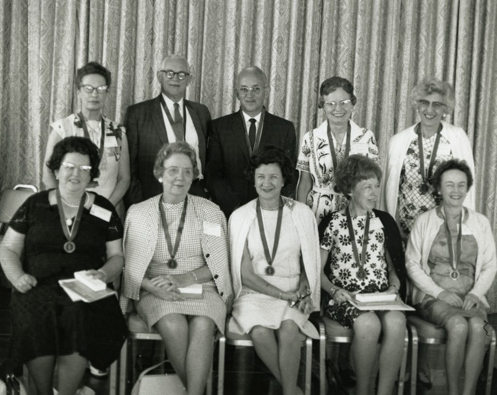 Black and white photo of 8 women and 2 men posing shoulder to shoulder, wearing medals.