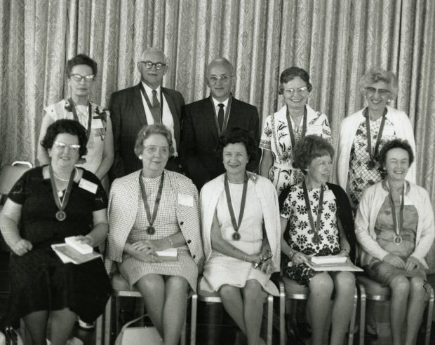 Black and white photo of 8 women and 2 men posing shoulder to shoulder, wearing medals.