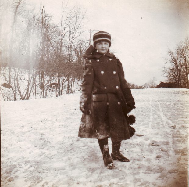 Black and white photograph of a child in winterwear posing on a snow trodden path.