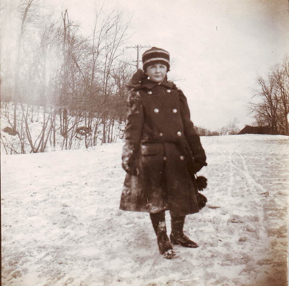 Black and white photograph of a child in winterwear posing on a snow trodden path.