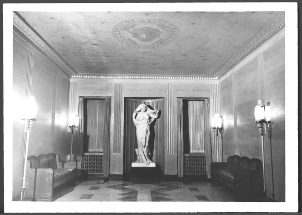 Black and white image of a tiled lobby with benches on the right and left sides, floor lamps, and a white marble statue at the centre.