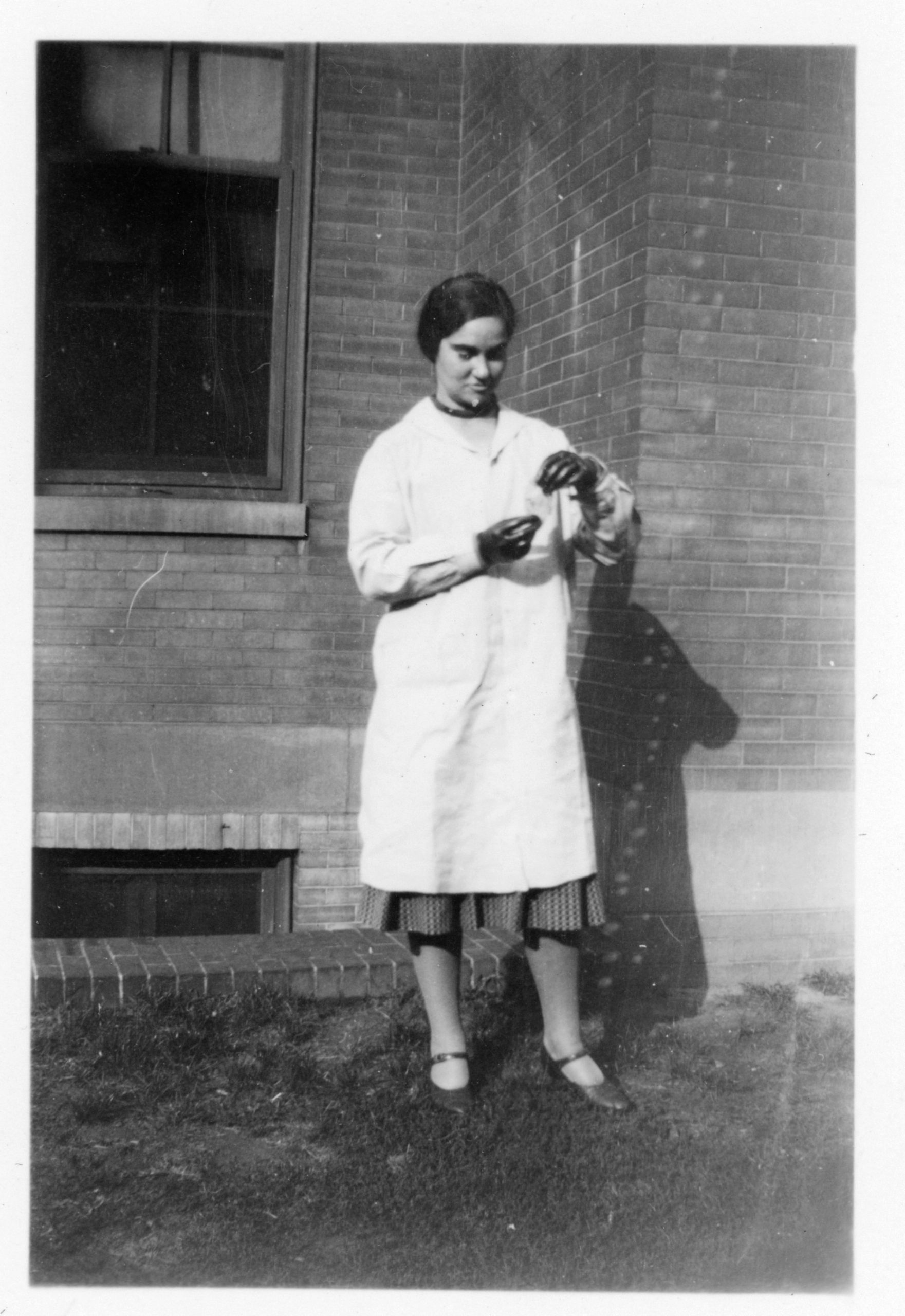 Black and white photo of a woman wearing a white labcoat and black rubber gloves, staring down at a small piece of paper she is holding between her hands.
