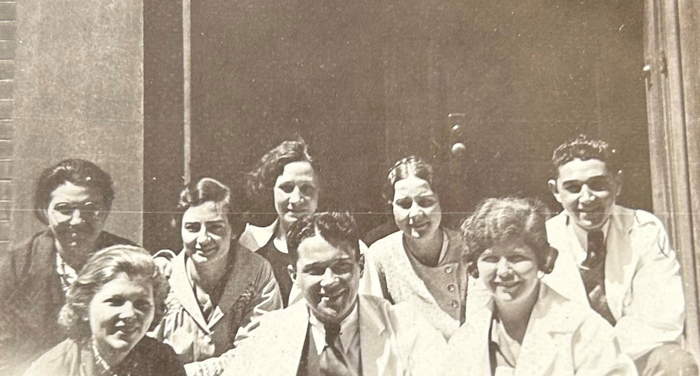 Black and white group photo of students of all genders posing in two rows, wearing lab coats. Photo is cropped above the shoulders.