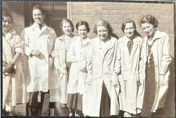 Black and white photo of seven classmates posing in a row, leaning against a building and wearing their lab coats.