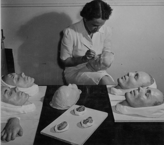 Black and white photo of a woman painting a moulage of a face. Various other medical moulages are sitting on the table in front of her, including more faces, a hand and ears.
