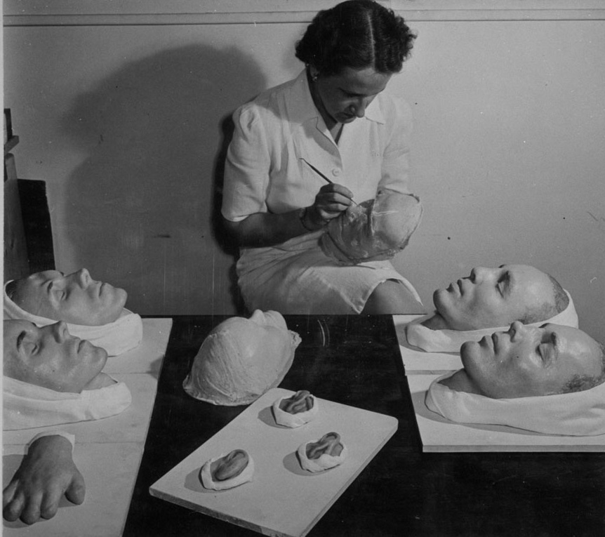 Black and white photo of a woman painting a moulage of a face. Various other medical moulages are sitting on the table in front of her, including more faces, a hand and ears.