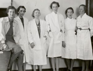 Photo en noir et blanc de six femmes posant côte à côte, portant des blouses de laboratoire.