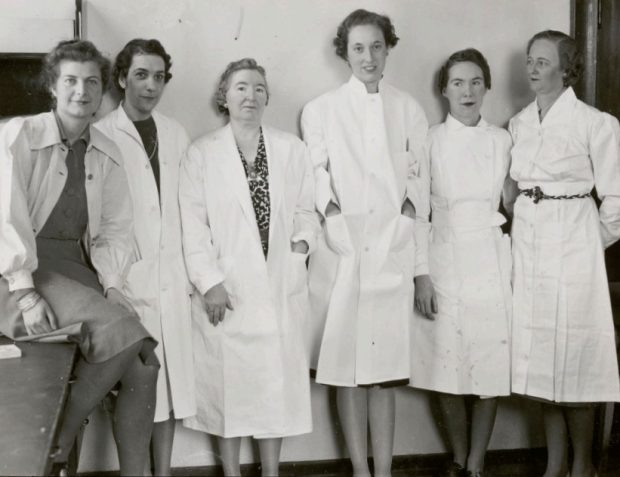 Black and white photo of 6 women posing shoulder to shoulder wearing lab coats.