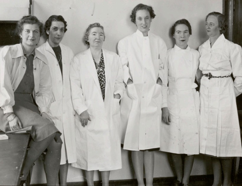 Black and white photo of 6 women posing shoulder to shoulder wearing lab coats.