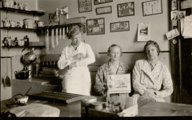Black and white photo of three women seated in a laboratory. In the left corner is a sink with shelves covered with cups, a kettle and a teapot. The rear wall is covered with framed photos of staff members.