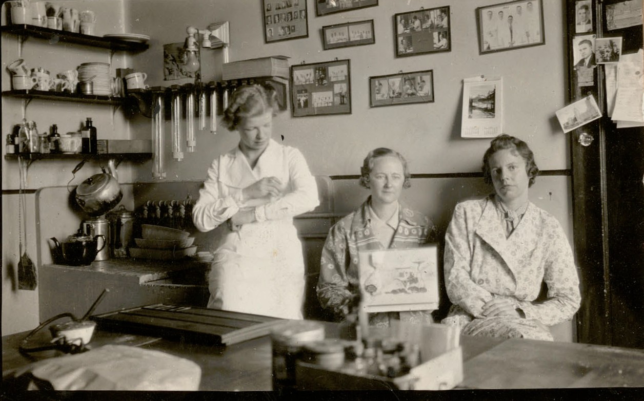 Black and white photo of three women seated in a laboratory. In the left corner is a sink with shelves covered with cups, a kettle and a teapot. The rear wall is covered with framed photos of staff members.
