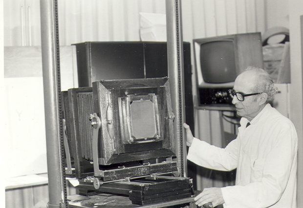 Black and white photo of an elderly photographer manipulating a large camera with audio-visual equipment in the background.