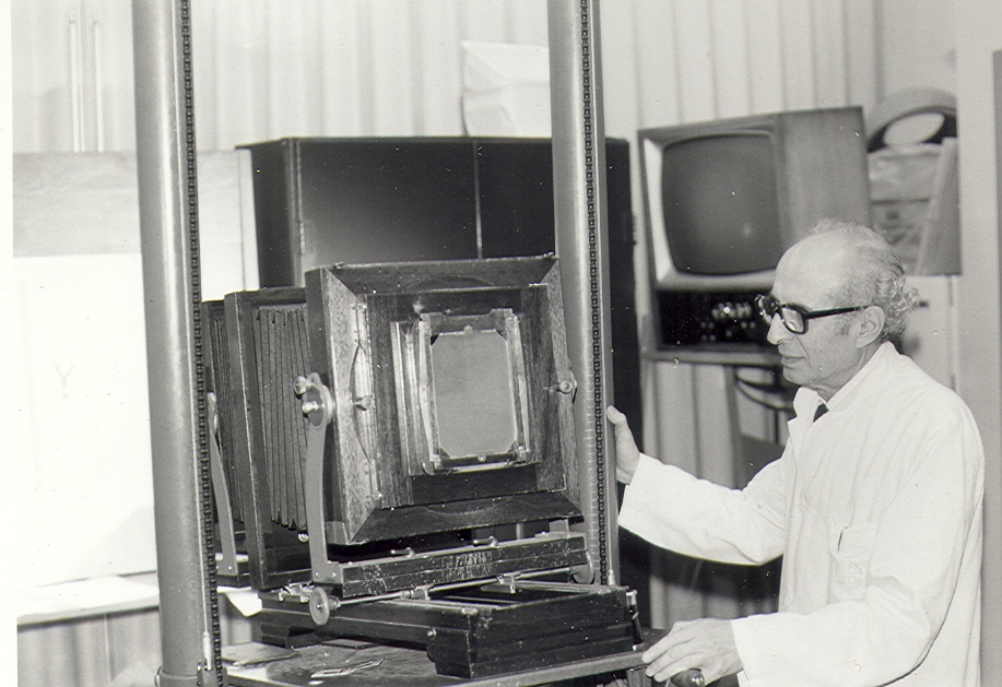 Black and white photo of an elderly photographer manipulating a large camera with audio-visual equipment in the background.