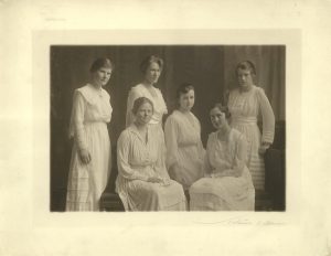 Photographie aux tons sépia montrant six femmes vêtues de robes blanches, posant et regardant l'appareil. Trois sont assises au centre, les trois autres debout.