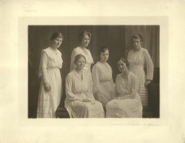 Sepia-toned photograph of six women wearing white dresses and posing and looking at the camera. Three are seated in the centre of the group, and three are standing.