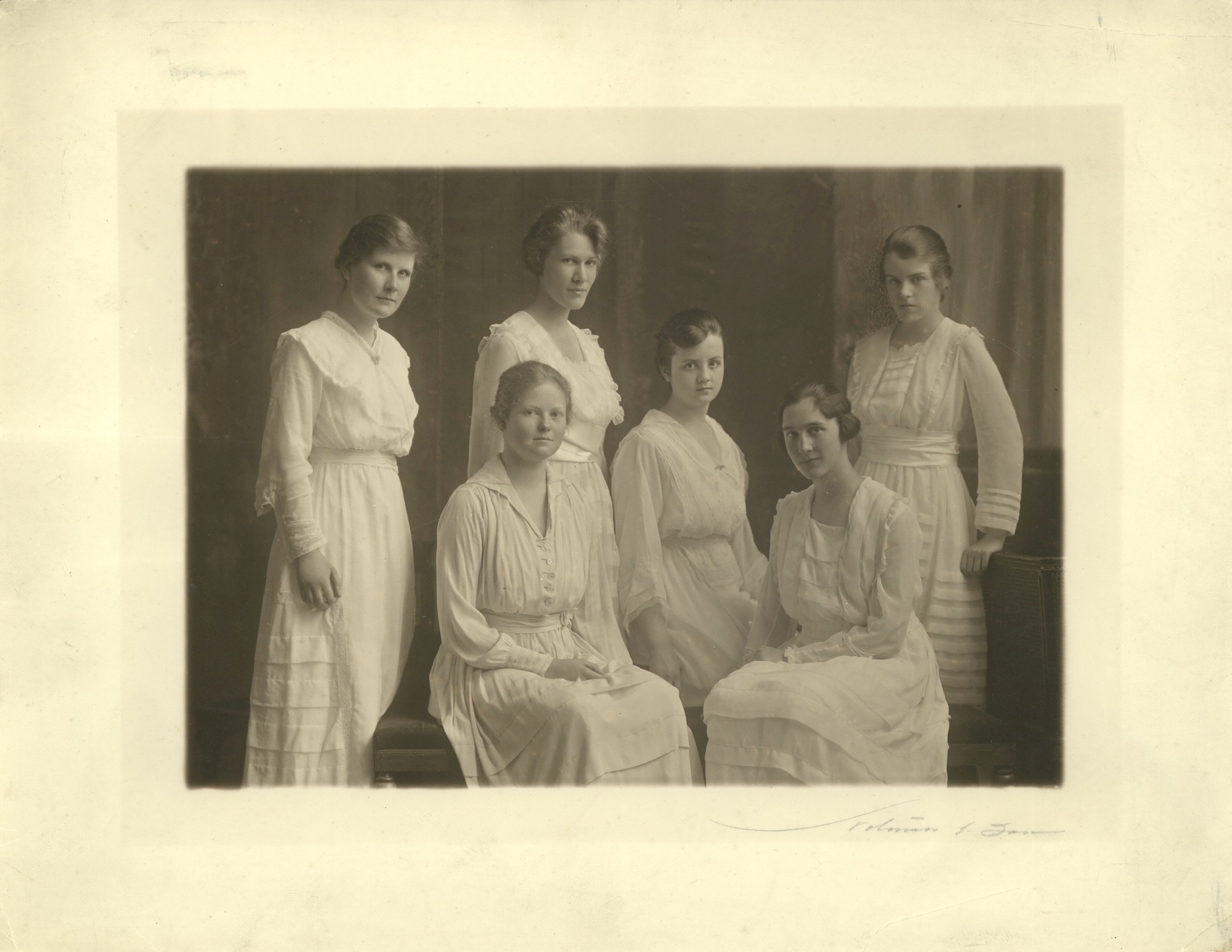 Sepia-toned photograph of six women wearing white dresses and posing and looking at the camera. Three are seated in the centre of the group, and three are standing.