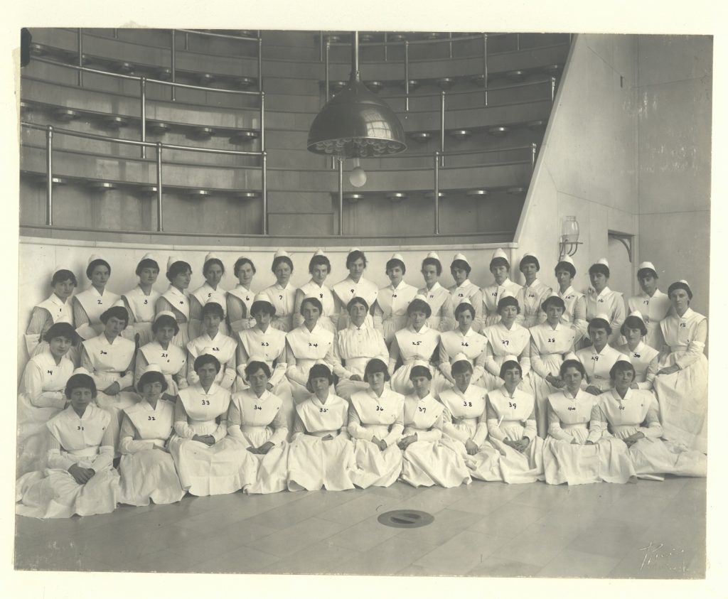 Black and white photo of a group of nursing graduates in white uniforms, posing in an operating theatre.