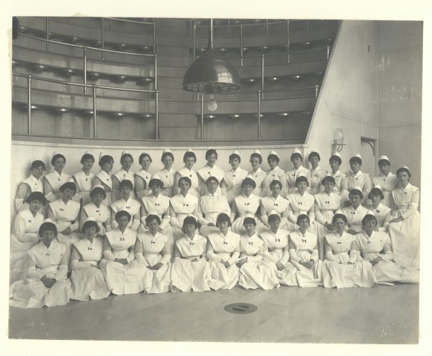 Black and white photo of a group of nursing graduates in white uniforms, posing in an operating theatre.