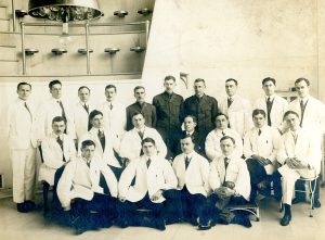 Twenty-one men posing in three rows in an operating theatre of a hospital, wearing white lab coats or military uniforms.