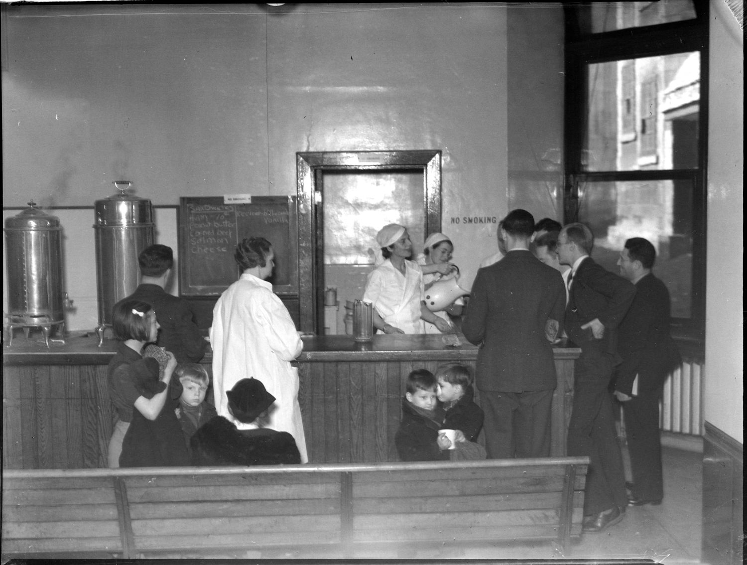 Black and white photograph of various people of all ages being served from behind a cafeteria counter. Two servers wear white uniforms; one pours from a pitcher.