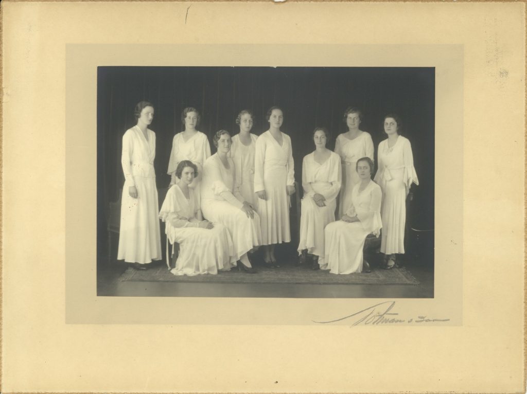 Sepia photo of 10 young women dressed in white gowns, some sitting and some standing.