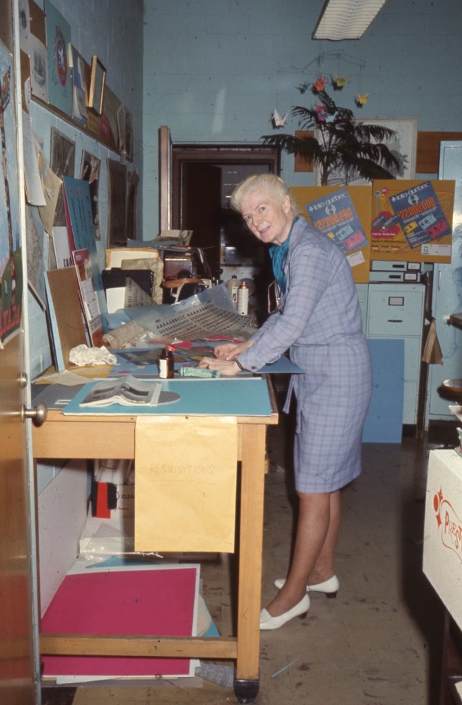 Colour photo of Mary Gzowski in her studio standing over her work desk with various projects and posters on her desk and walls.
