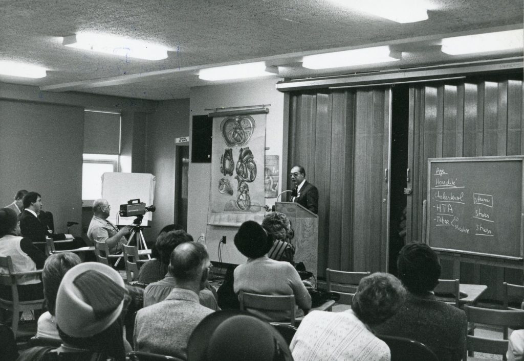 Black and white photo of a doctor giving a presentation to people seated at desks, using a medical illustration by Pauline Larivière.
