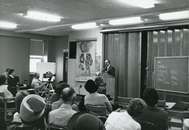 Black and white photo of a doctor giving a presentation to people seated at desks, using a medical illustration by Pauline Larivière.