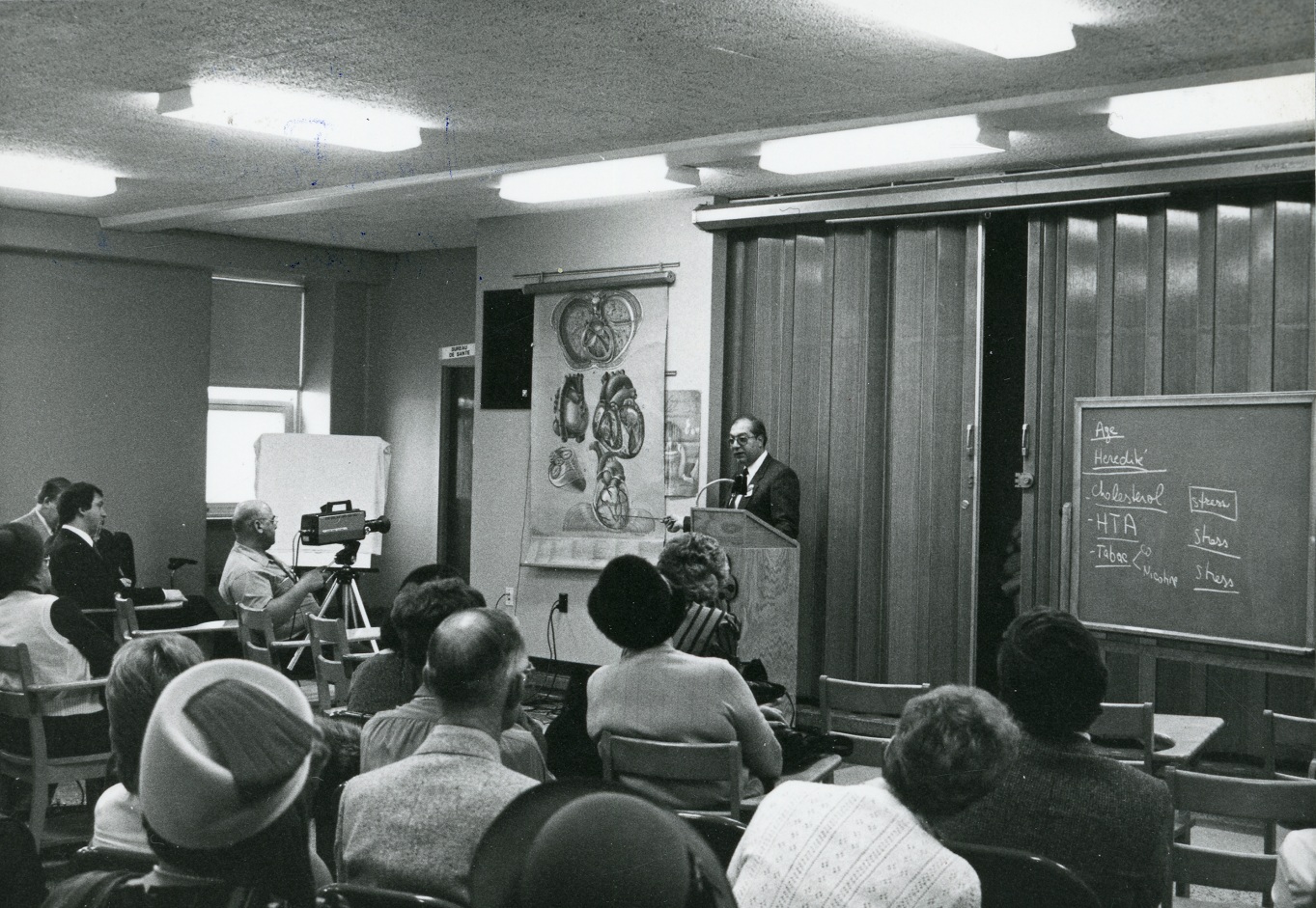 Black and white photo of a doctor giving a presentation to people seated at desks, using a medical illustration by Pauline Larivière.