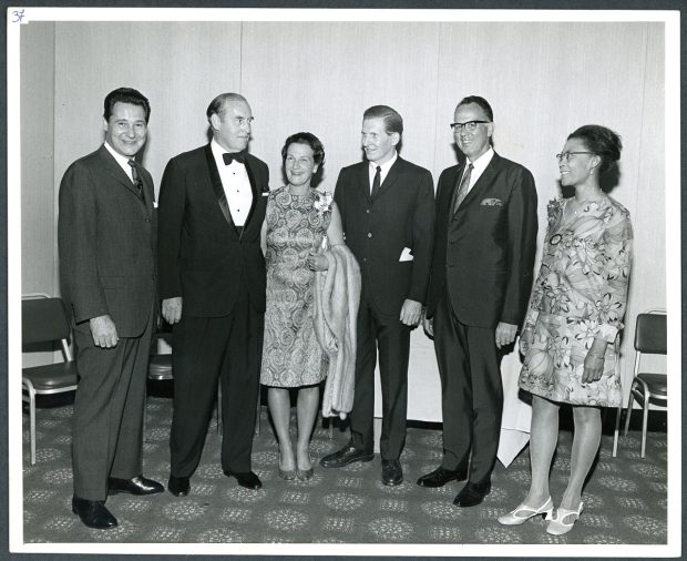 Black and white photo of four men and two women posing shoulder to shoulder, dressed in formal attire.