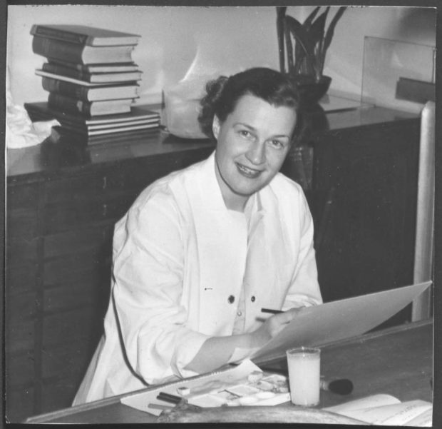 Black and white photo of a woman sitting at a desk, sketching on illustration board.