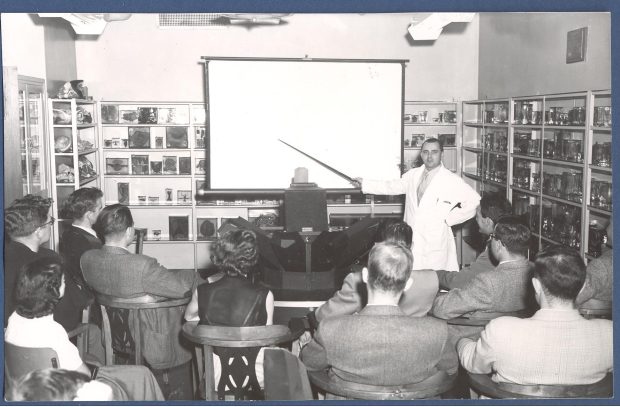 Black and white photograph of a classroom lined with medical teaching slides and specimens. A man in a white coat faces the camera and uses a pointer to indicate to students.