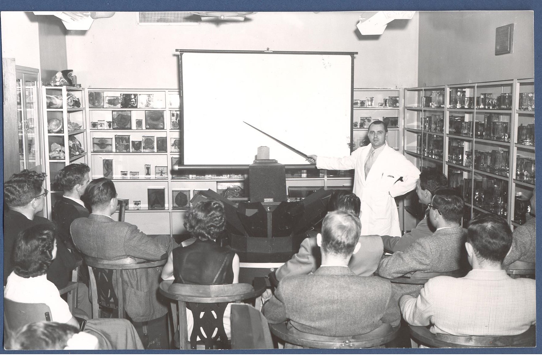 Black and white photograph of a classroom lined with medical teaching slides and specimens. A man in a white coat faces the camera and uses a pointer to indicate to students.