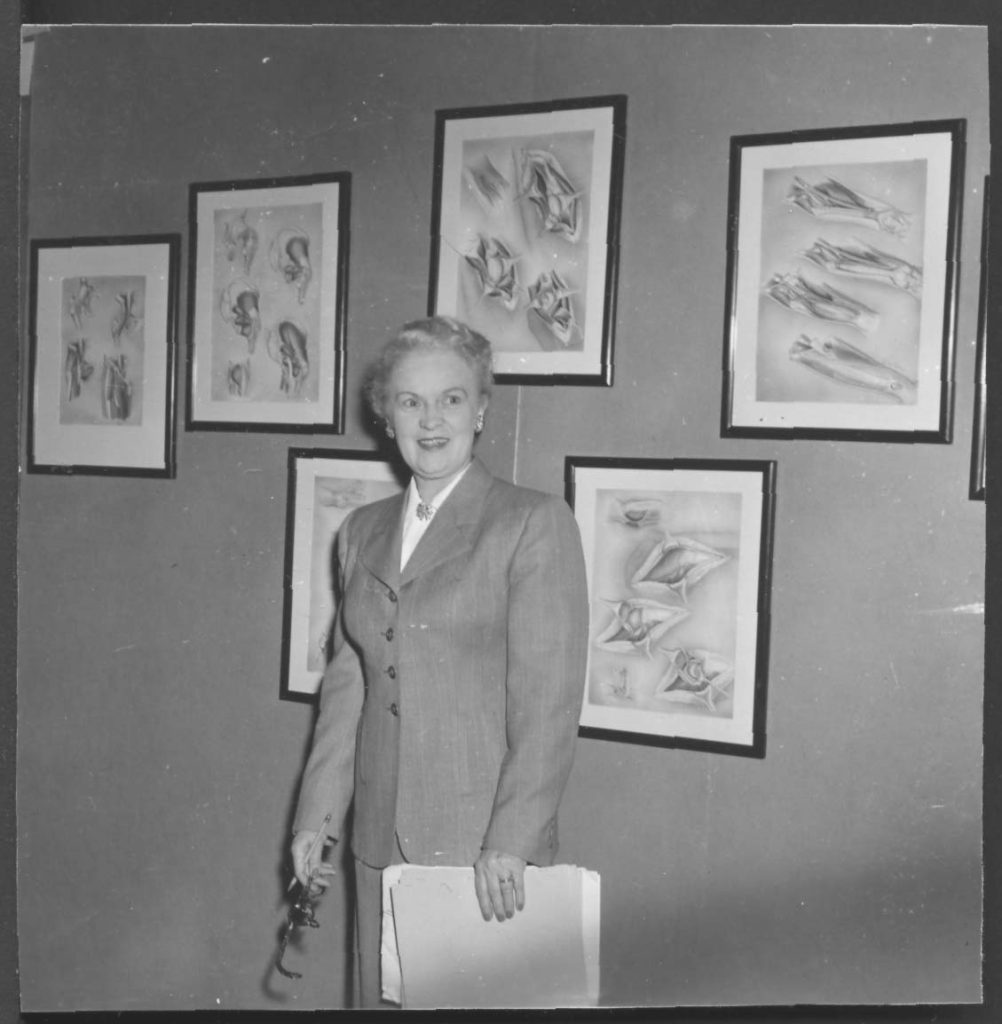Black and white photo of Winifred Rock standing in front of illustrations by Helen MacArthur.