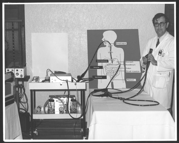 Black and white photo of a doctor setting up a medical teaching display featuring an illustration of a human body connected to various tubes and medical equipment.