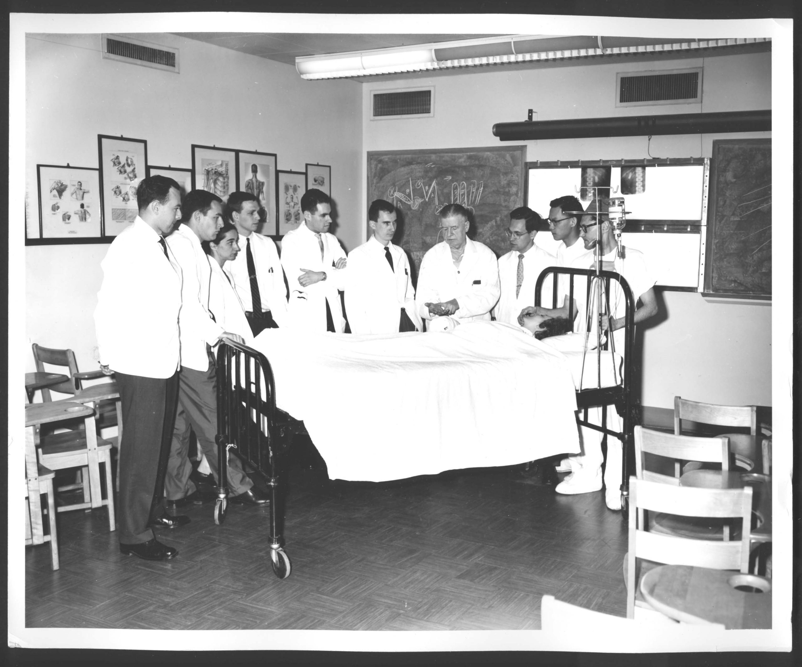 Black and white photo of a doctor teaching medical residents at a patient's bedside. Medical illustrations can be seen hanging in the background.