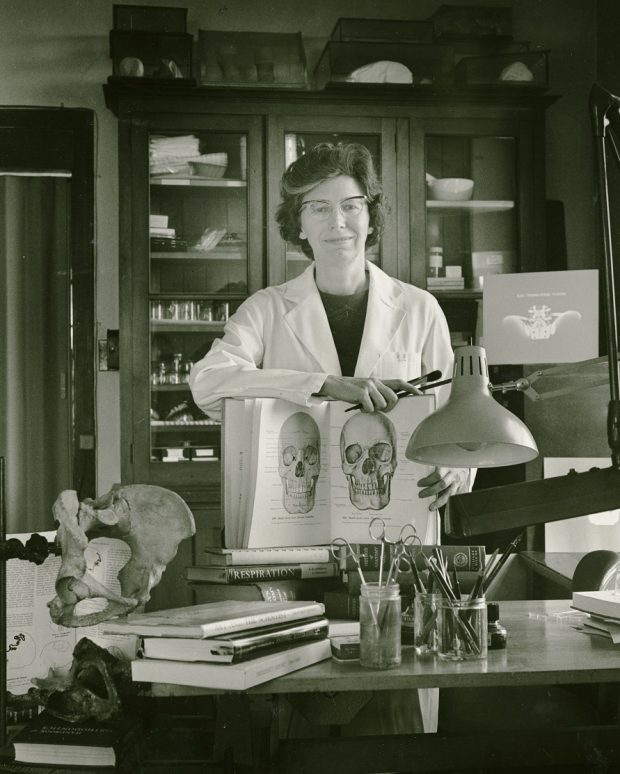 Black and white photo of a woman wearing a lab coat in a laboratory, displaying an open book of medical illustrations.