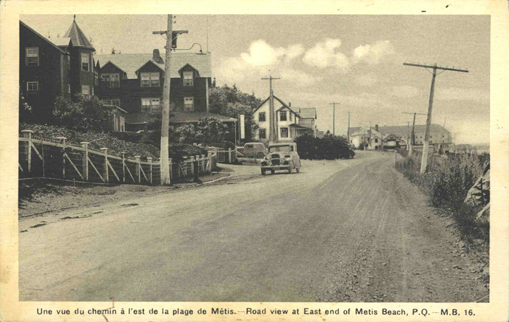 Black and white postcard printed on card presents the Chemin du Roi. The dirt road crossing the town of Métis-sur-Mer. A car is parked along the road. Along the way there are 2 of the major hotels of the time as well as some of the magnificent summer residences.