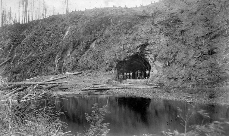 Portrait of 8 workers in the entrance of the diversion tunnel for the Tartigou River. The entrance to the tunnel is located at the bottom of a steep slope that has been entirely cleared, the stumps are still visible. A few meters down the tunnel entrance flows the river Tartigou River.