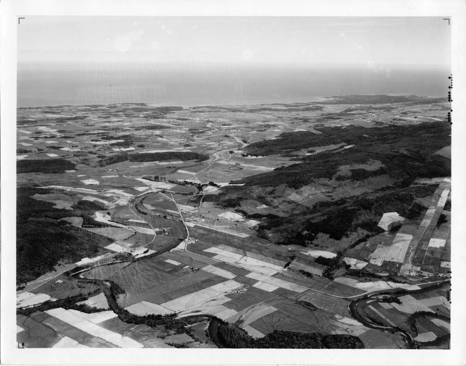 Oblique black and white aerial photograph showing agricultural and forested lands along the Mitis River to the St. Lawrence River. The different types of culture are presented as a checkerboard of different colours and textures.